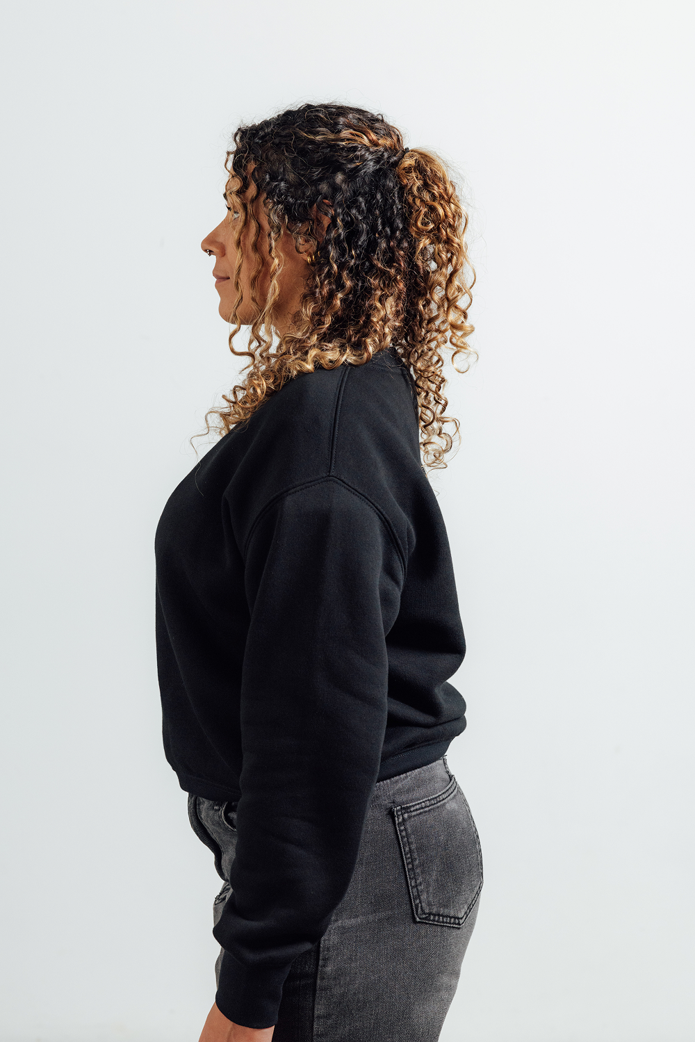 A woman shown in side profile wearing a black crewneck sweatshirt and dark gray jeans against a plain light gray studio background.