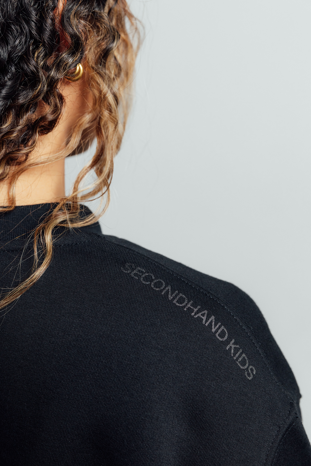 Close-up back view of a person wearing a black sweatshirt with 'SECONDHAND KIDS' embroidered on the shoulder, against a light gray background.