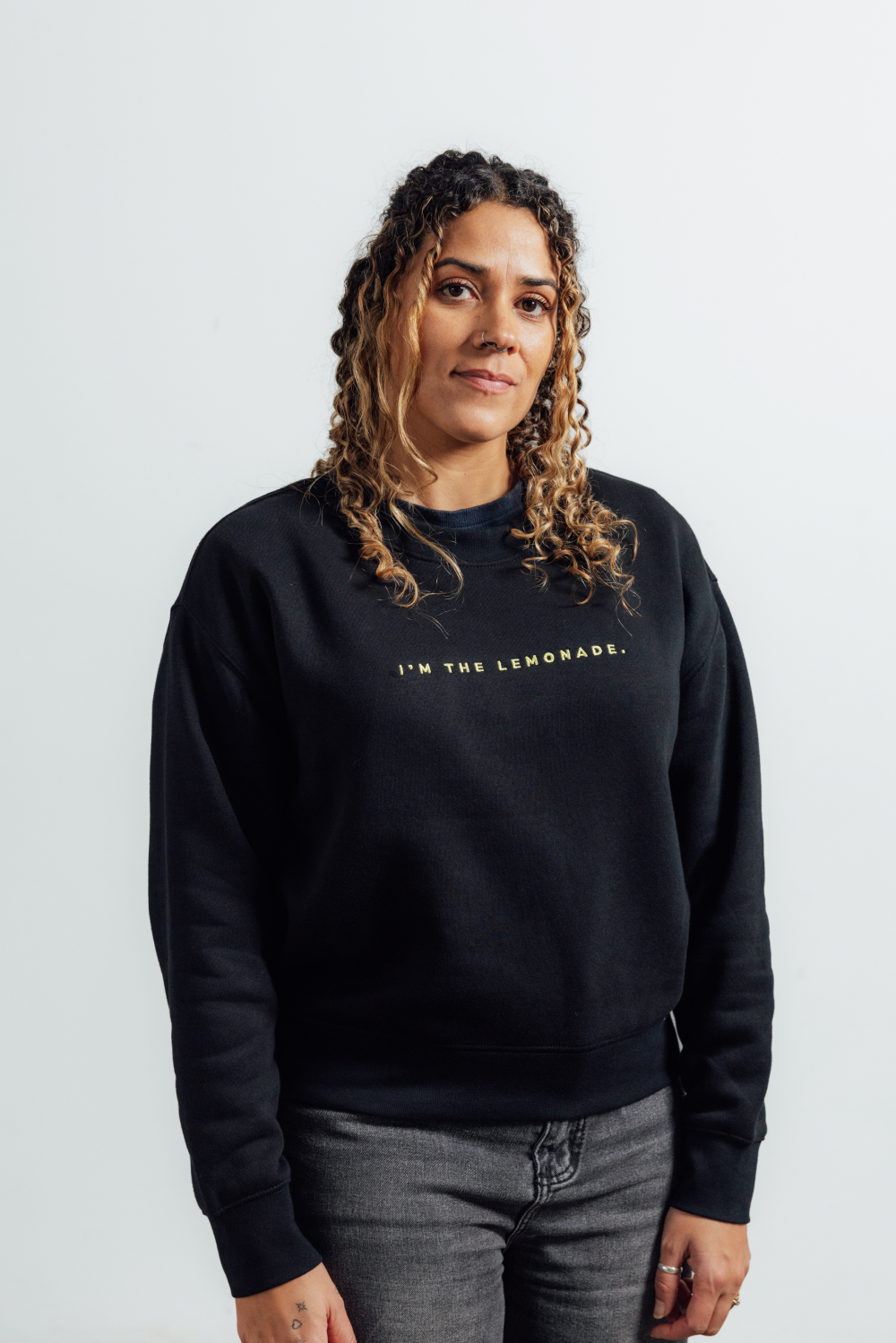A woman with curly brown hair wearing a black crewneck sweatshirt with the text 'I'M THE LEMONADE.' printed in gold across the chest, paired with gray jeans, standing against a light gray background.