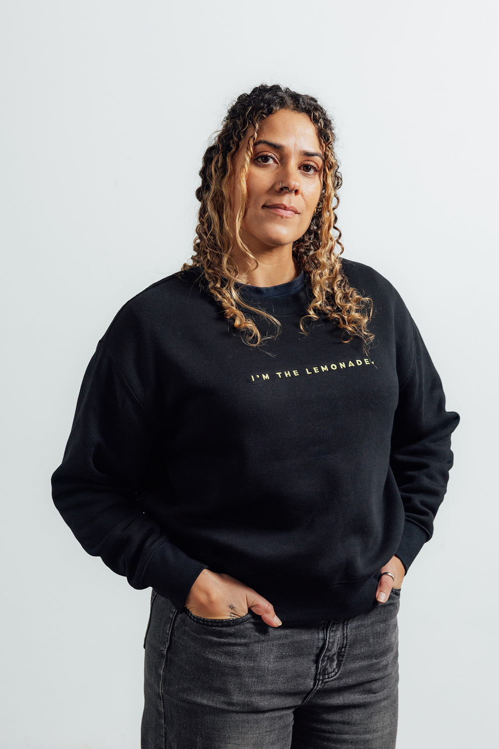 A woman with curly brown hair wearing a black crewneck sweatshirt with gold text reading 'I'M THE LEMONADE.' across the chest, paired with dark gray jeans, standing against a light gray studio background.