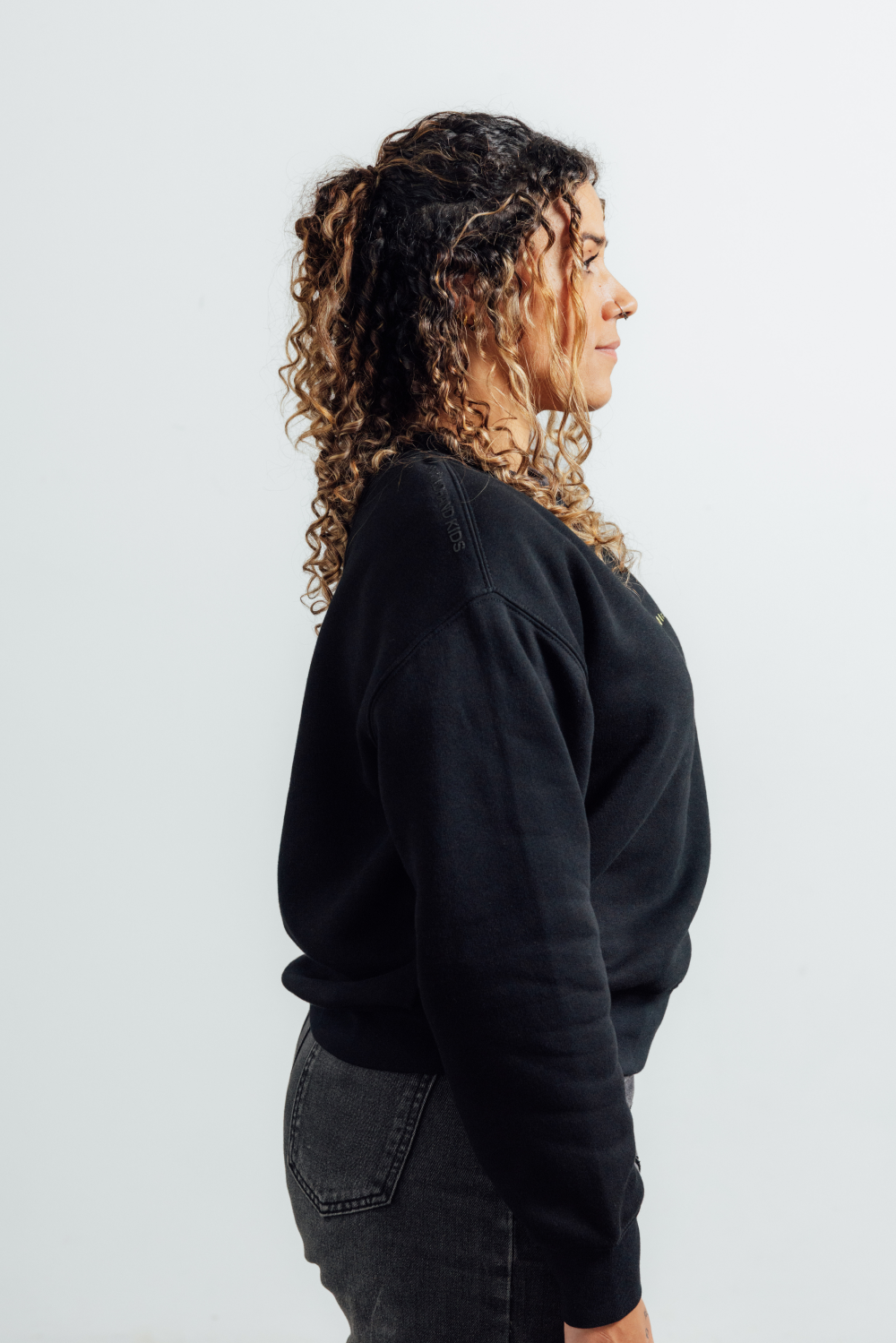 A woman with curly brown and blonde hair is shown in profile wearing a black crewneck sweatshirt and dark gray jeans against a plain light gray background.