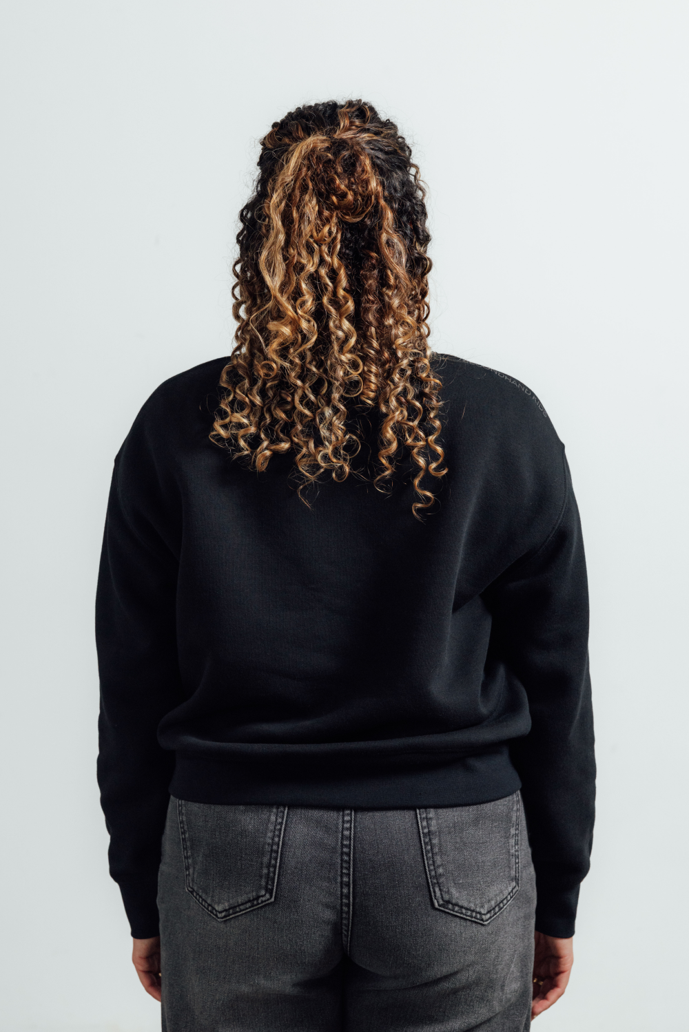 Back view of a person with curly brown and blonde hair wearing a black crewneck sweatshirt and dark gray jeans against a light gray background.