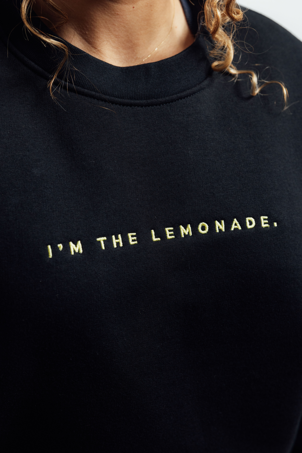 Close-up of a person wearing a black crewneck sweatshirt with the phrase 'I'M THE LEMONADE.' printed in gold across the chest.