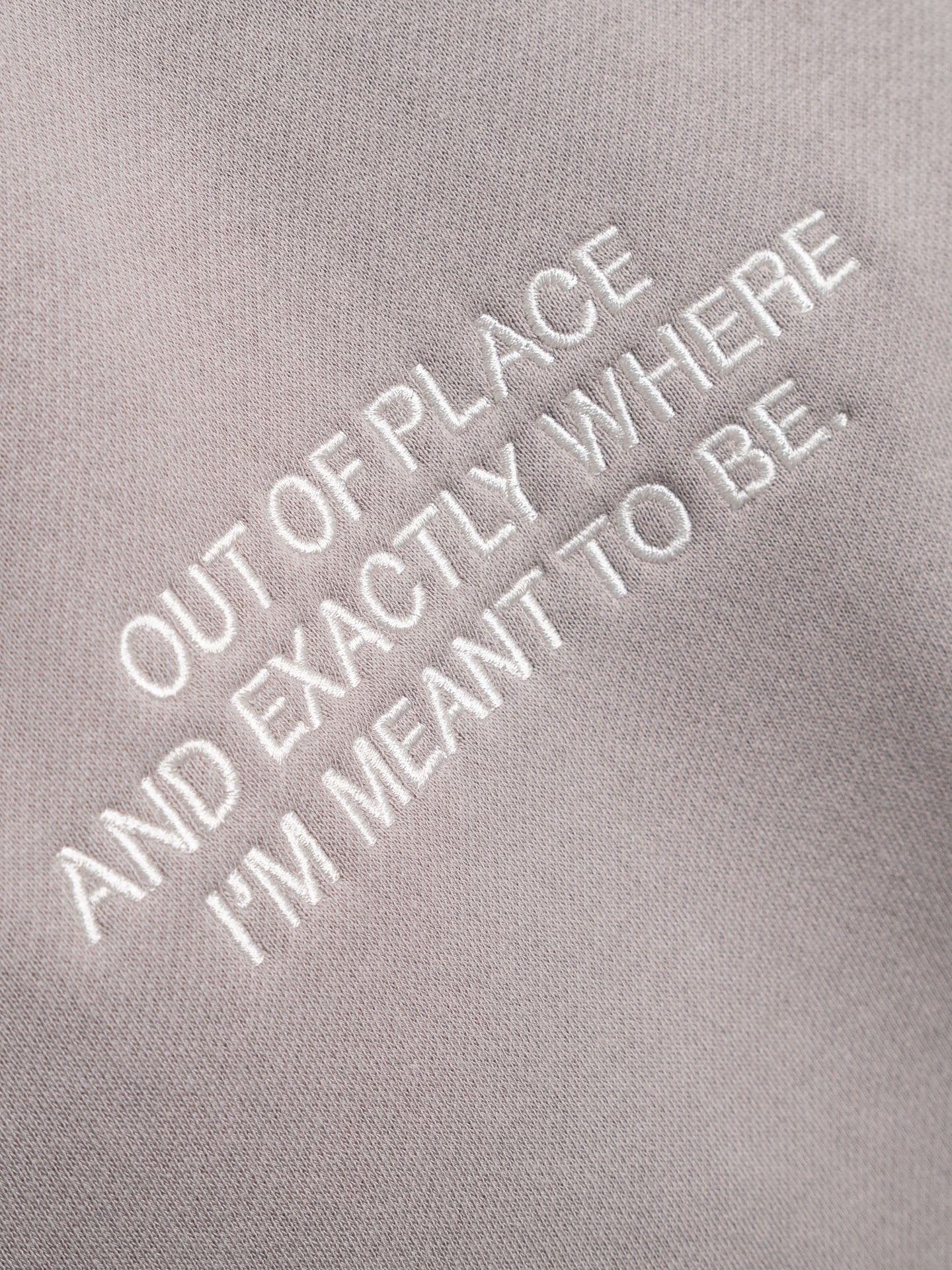 Embroidered text on a gray background reading 'OUT OF PLACE AND EXACTLY WHERE I'M MEANT TO BE'.