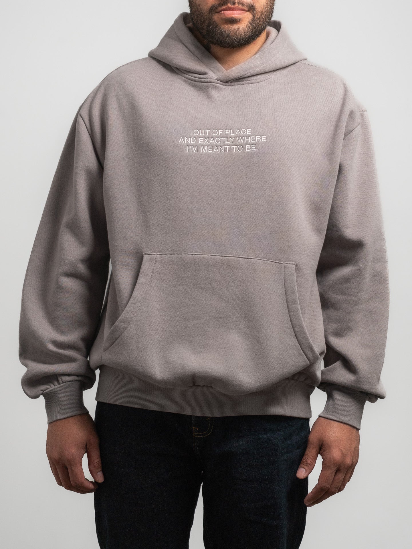 Male wearing a grey SecondHand Kids 'Out of Place ' hoodie with embroidered text reading "Out of place, and exactly where I’m meant to be". Standing against a plain background.