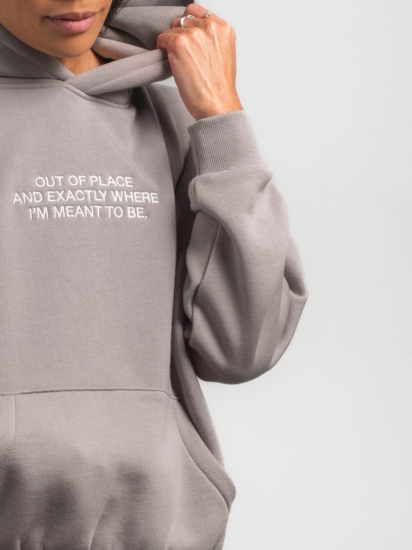 Gray hoodie with text 'OUT OF PLACE AND EXACTLY WHERE I'M MEANT TO BE.' on a light gray background