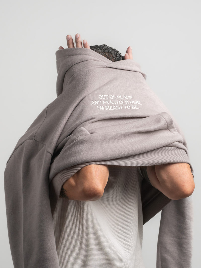 Person wearing a grey SecondHand Kids hoodie with text reading ‘Out of place, and exactly where I’m meant to be.’”, standing against a plain background.